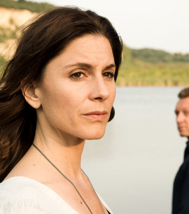 "In Wahrheit - Still ruht der See": Judith Mohn (Christina Hecke) blickt nachdenklich aus dem Bild. Hinter ihr steht Freddy Breyer (Robin Sondermann) und schaut zu Judith hin. Im Hintergrund ist ein See zu sehen.