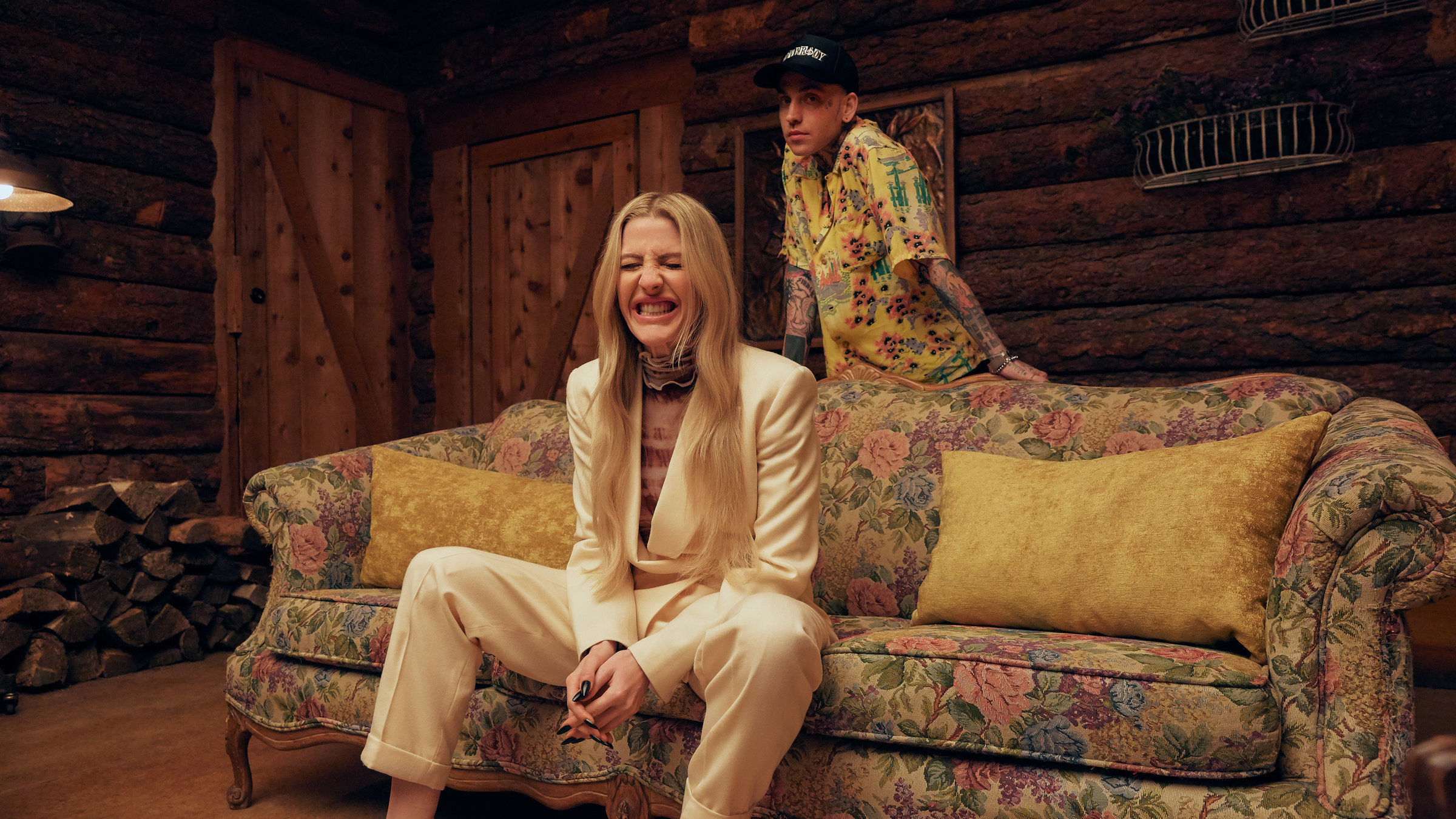 "Ellie Goulding: Live at the Victoria & Albert Museum": Auf einem altmodischen Sofa mit Blumenmuster sitzt eine Frau mit langen blonden Haaren in einem weißen Hosenanzug. Sie grinst und kneift die Augen zusammen. Hinter ihr steht ein junger Mann in gemustertem Hemd mit Baseballcap.