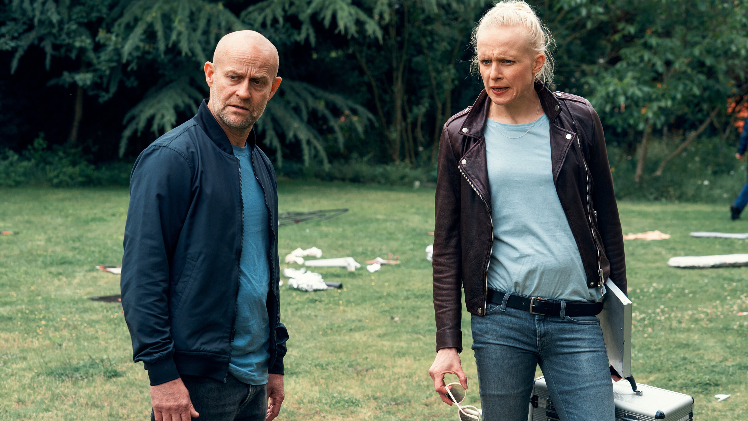 "Jenseits der Spree - Invasion": Robert (Jürgen Vogel) steht gemeinsam mit Dr. Schäfer (Anne Müller) auf einer Wiese. Überall liegt Müll. Dr. Schäfer hält einen silbernen Koffer in der Hand und hat ein Tablett unter den Arm geklemmt. Beide schauen zur Seite.