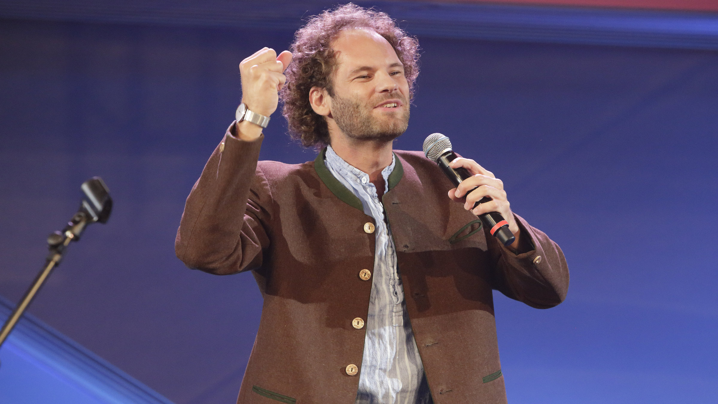 "Maxi Schafroth: Faszination Bayern": Maxi Schafroth steht auf der Bühne mit einem Mikrofon in der linken Hand und trägt eine braune Trachtenjacke.