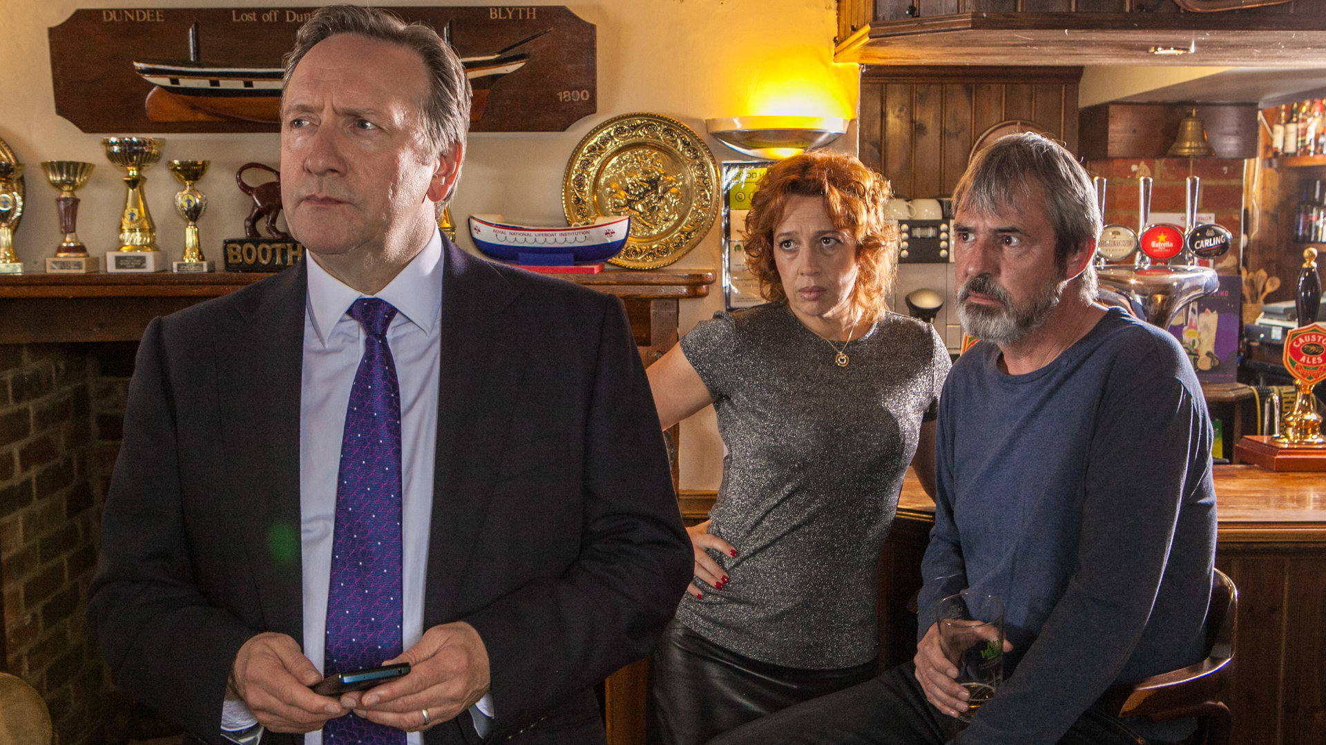 "Inspector Barnaby - Mord in bester Absicht":  Barnaby (Neil Dudgeon) hat sich soeben von den Wirtsleuten Lena Ferrera (Katy Cavanagh) und Mitch McAllister (Neil Morrissey) abgewandt, die im Hintergrund auf Barhockern an einer Theke sitzen und ihm betroffen in den Rücken schauen.