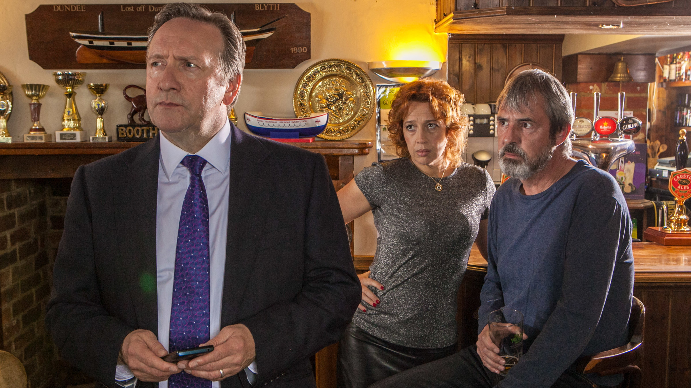 "Inspector Barnaby - Mord in bester Absicht":  Barnaby (Neil Dudgeon) hat sich soeben von den Wirtsleuten Lena Ferrera (Katy Cavanagh) und Mitch McAllister (Neil Morrissey) abgewandt, die im Hintergrund auf Barhockern an einer Theke sitzen und ihm betroffen in den Rücken schauen.