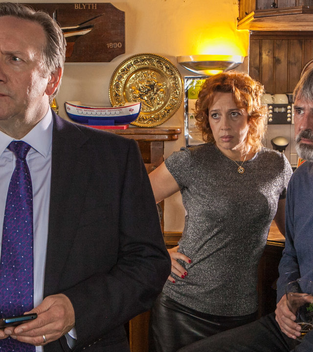 "Inspector Barnaby - Mord in bester Absicht":  Barnaby (Neil Dudgeon) hat sich soeben von den Wirtsleuten Lena Ferrera (Katy Cavanagh) und Mitch McAllister (Neil Morrissey) abgewandt, die im Hintergrund auf Barhockern an einer Theke sitzen und ihm betroffen in den Rücken schauen.