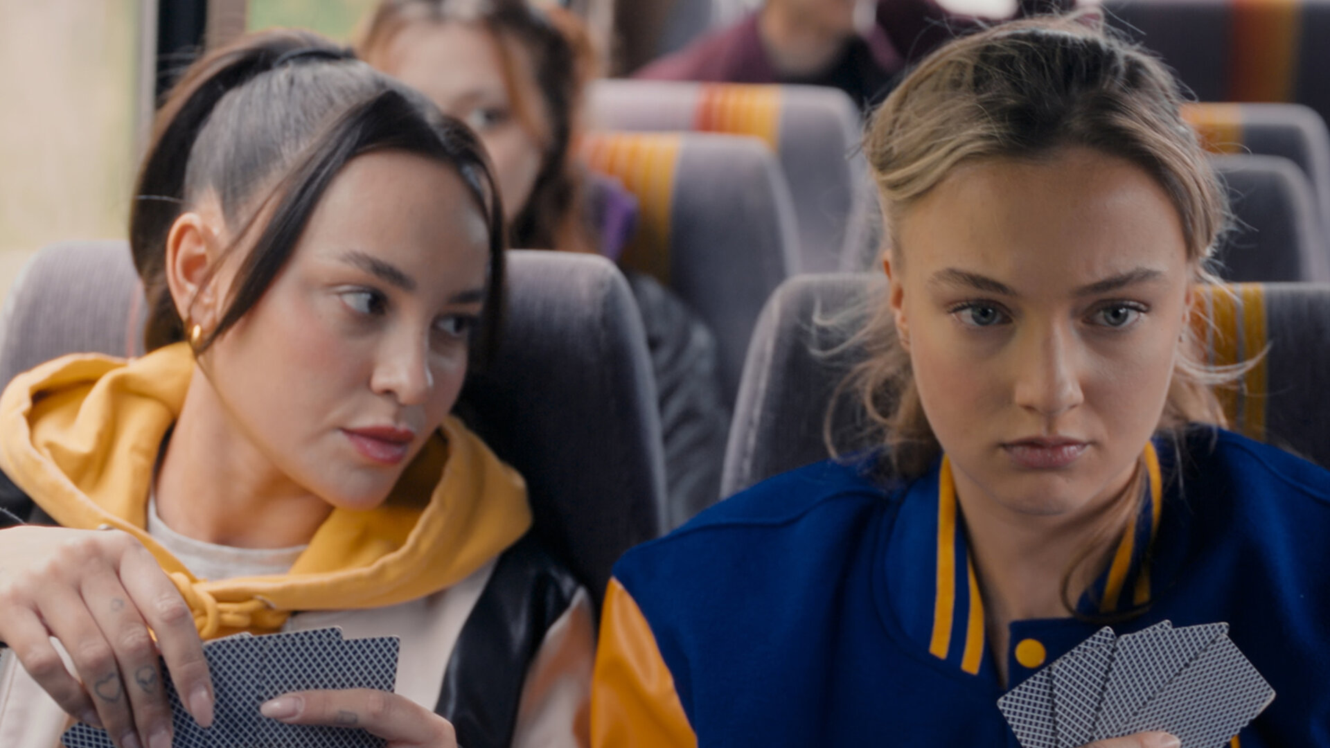 "Club der Dinosaurier - Das Camp": Esra (Margarita Nigamtullin) und Fiona (Lilly Krug) sitzen in einem Bus. Sie sind umgeben von Teenagern (Komparsen) und spielen Karten.