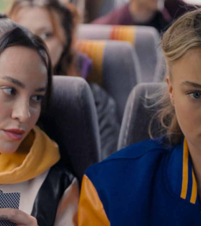 "Club der Dinosaurier - Das Camp": Esra (Margarita Nigamtullin) und Fiona (Lilly Krug) sitzen in einem Bus. Sie sind umgeben von Teenagern (Komparsen) und spielen Karten.