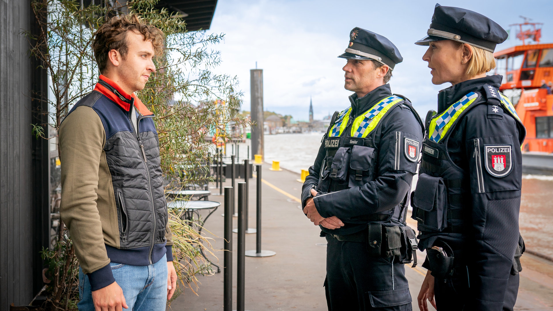 "Notruf Hafenkante - Funkstille": Lennard Böhm (Tim Bülow) steht vor seinem Bistro am Hafen. Ihm gegenüber stehen der Polizist Mattes Seeler (Matthias Schloo) und seine Kollegin Melanie Hansen (Sanna Englund). Beide sehen ihn an.