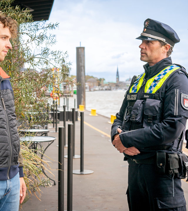 "Notruf Hafenkante - Funkstille": Lennard Böhm (Tim Bülow) steht vor seinem Bistro am Hafen. Ihm gegenüber stehen der Polizist Mattes Seeler (Matthias Schloo) und seine Kollegin Melanie Hansen (Sanna Englund). Beide sehen ihn an.