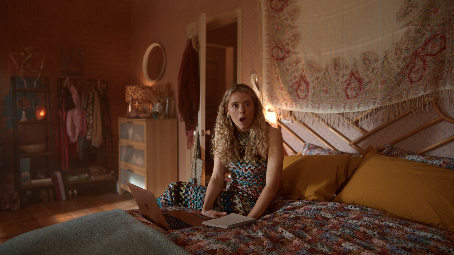"Extraordinary - Der Exorzismus der Carrie Jackson": Carrie (Sofia Oxenham) sitzt auf einem Bett in einem gemütlich eingerichteten Schlafzimmer, vor ihr liegen ein Laptop und ein Notizbuch. Mit offenem Mund blickt sie erstaunt zur Seite.