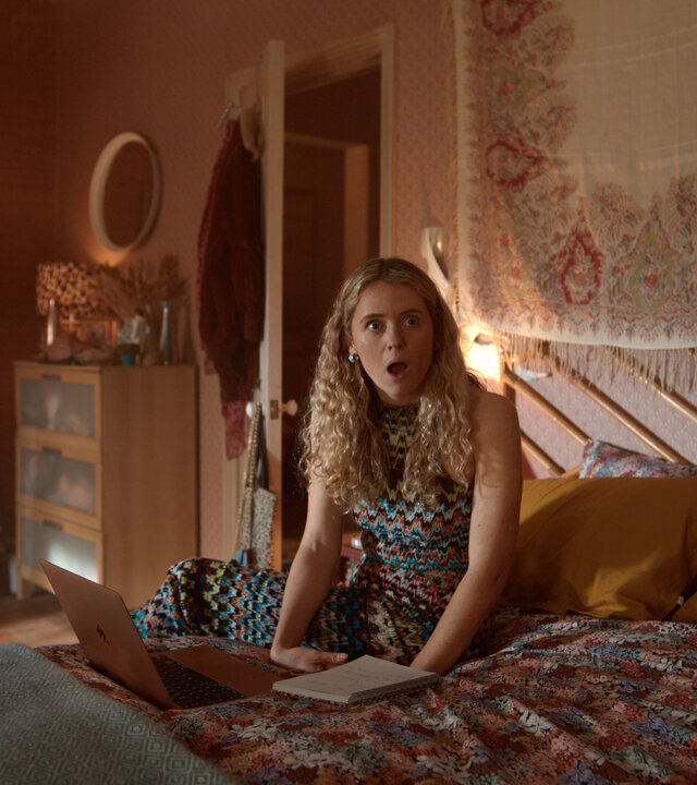 "Extraordinary - Der Exorzismus der Carrie Jackson": Carrie (Sofia Oxenham) sitzt auf einem Bett in einem gemütlich eingerichteten Schlafzimmer, vor ihr liegen ein Laptop und ein Notizbuch. Mit offenem Mund blickt sie erstaunt zur Seite.