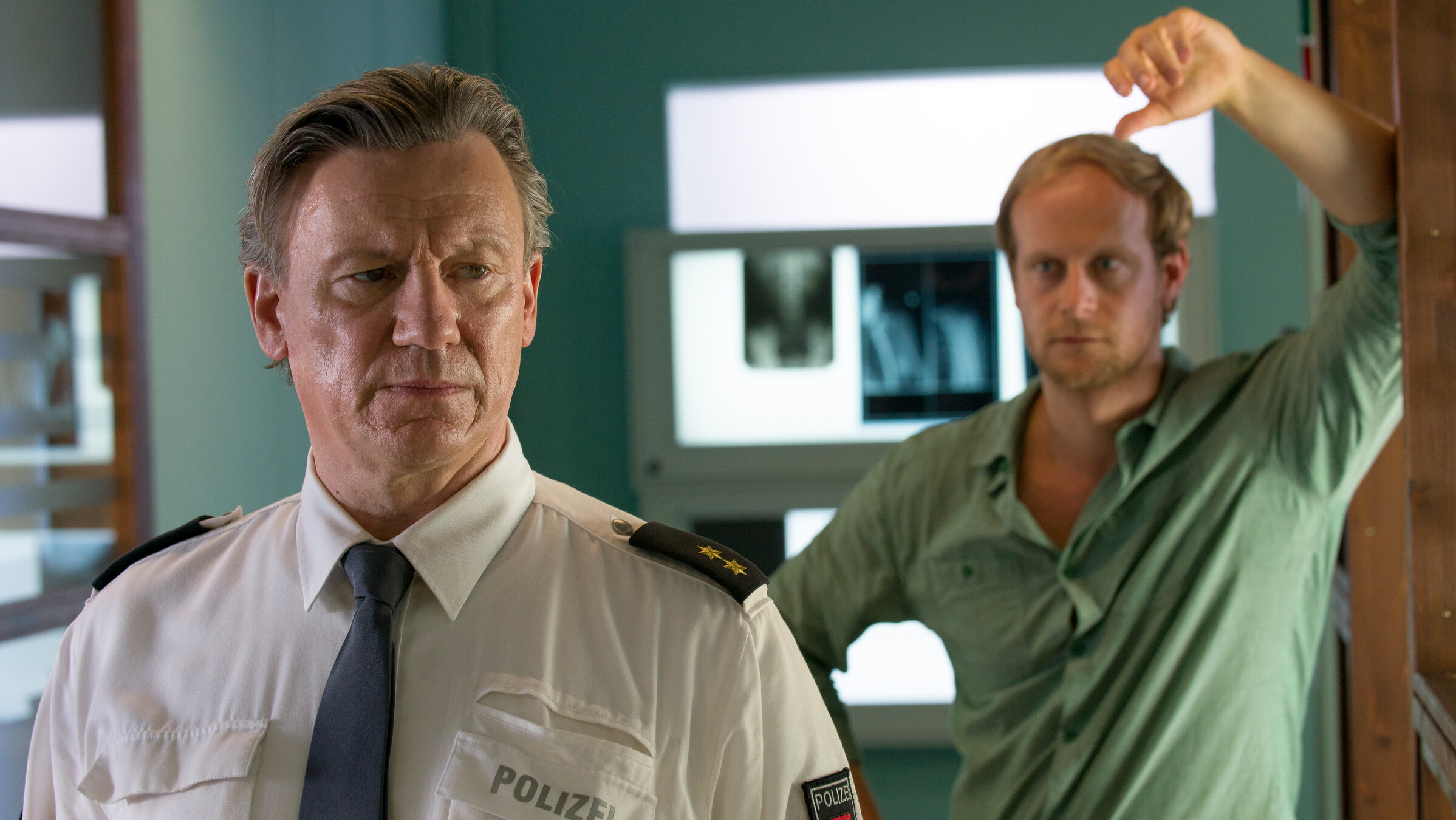 "Notruf Hafenkante - Einstand": Wolf Haller (Hannes Hellmann) steht nachdenklich im Büro. Hinter ihm steht der Journalist Elias Vancleef (Gabor Biedermann) und sieht in fragend an.
