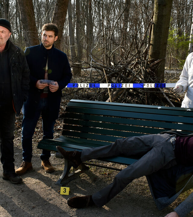 "Ein starkes Team - Für immer jung": Auf einer grünen Parkbank liegt ein Toter, Alexander Möwe (Vinzenz Wagner. Kleine gelbe Spurennummern sind um die Leiche verteilt. Dahinter steht Dr. Gabriele Simkeit (Eva Sixt) im weißen Overall und Handschuhen. Das Ermittlerteam aus Linett Wachow (Stefanie Stappenbeck), Otto Garber (Florian Martens) und Nils Makowski (Lucas Reiber) steht neben der Bank. Linett schaut auf den Toten, Otto und Nils schauen ihre Kollegin an. Im Hintergrund flattert zwischen kahlen Bäumen und Sträuchern ein blau-weißes Absperrband mit der Aufschrift "Polizei".