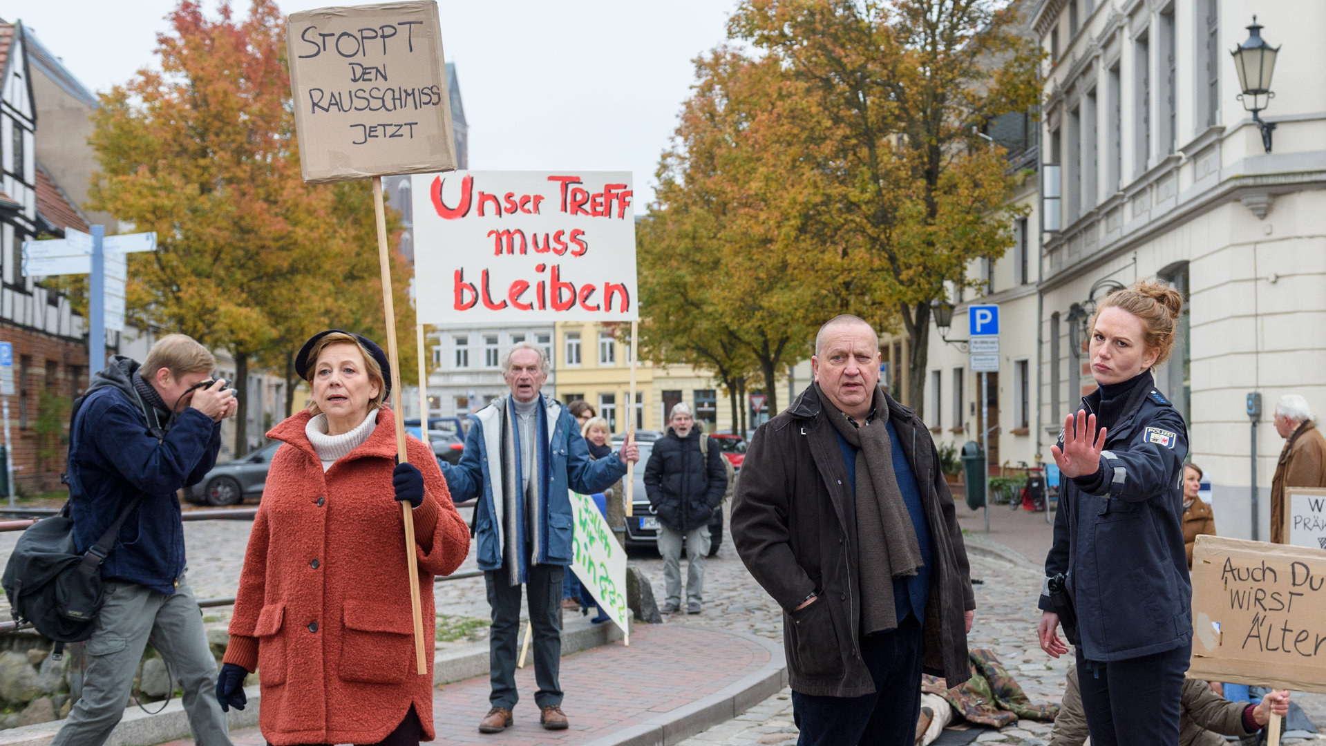 "SOKO Wismar – Hemingways Bauch": Auf einer Straße in der Wismarer Innenstadt blockieren Marlies Plattke (Judith von Radetzky) und weitere Demonstrierende die Durchfahrt. Paula Moorkamp (Stella Hinrichs) und Jan Reuter (Udo Kroschwald) stehen auf der Straße. Holger Nowak (Till Demuth) fotografiert die Demonstration.
