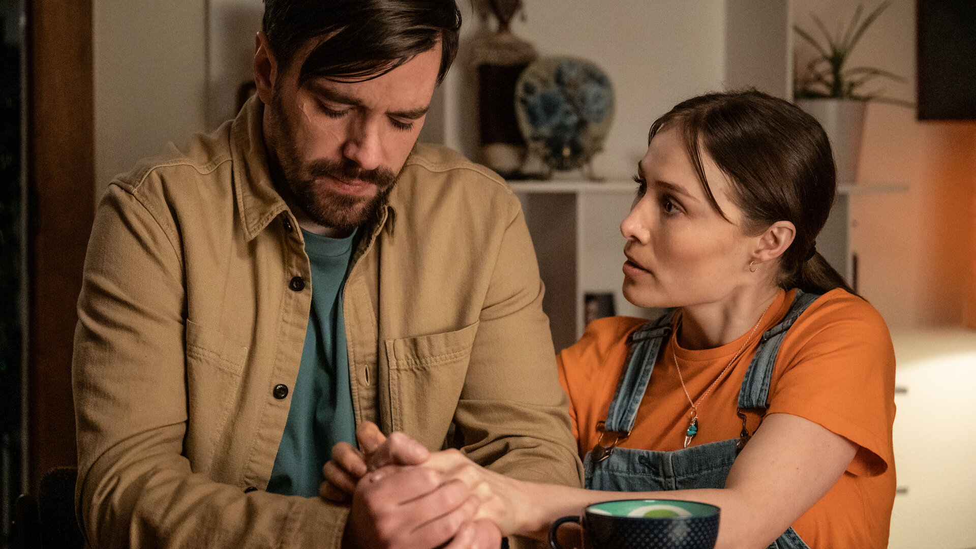 "Blind Sherlock - Undercover": Roman (Bart Kelchtermans) sitzt in einem Raum. Er wirkt traurig. Neben ihm kniet Caro (Sigrid ten Napel) und hält seine Hände. Vor ihnen auf dem Tisch steht eine Tasse.