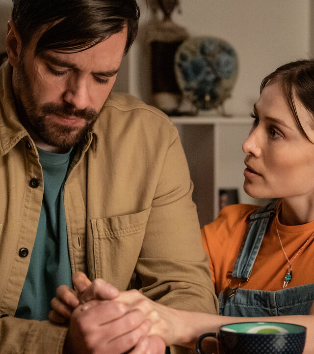 "Blind Sherlock - Undercover": Roman (Bart Kelchtermans) sitzt in einem Raum. Er wirkt traurig. Neben ihm kniet Caro (Sigrid ten Napel) und hält seine Hände. Vor ihnen auf dem Tisch steht eine Tasse.