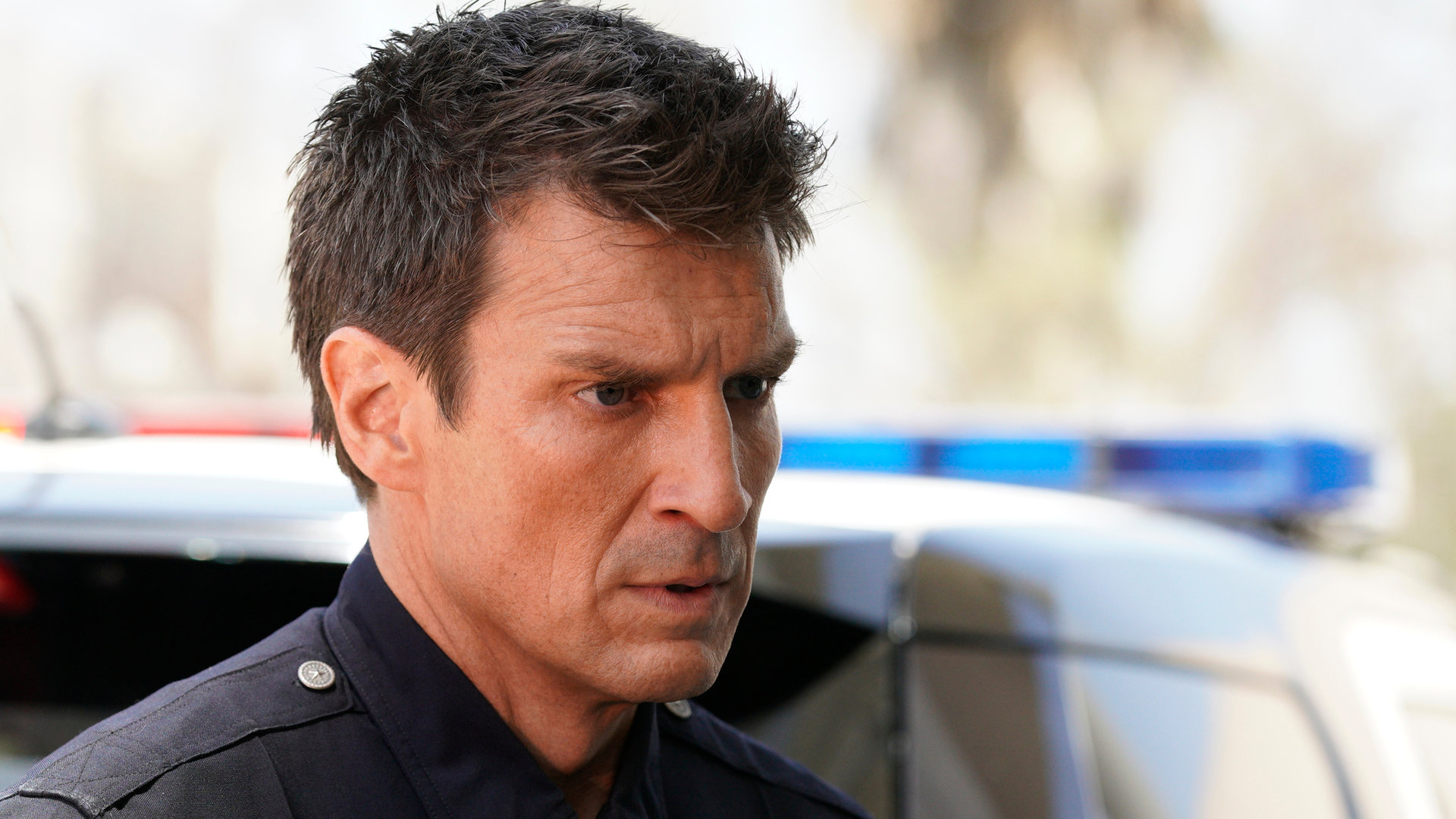 "The Rookie - Nachtschatten": John Nolan (Nathan Fillion) schaut mit entschlossenem Blick in die Ferne. Im Hintergrund ist ein Polizeiauto zu sehen.