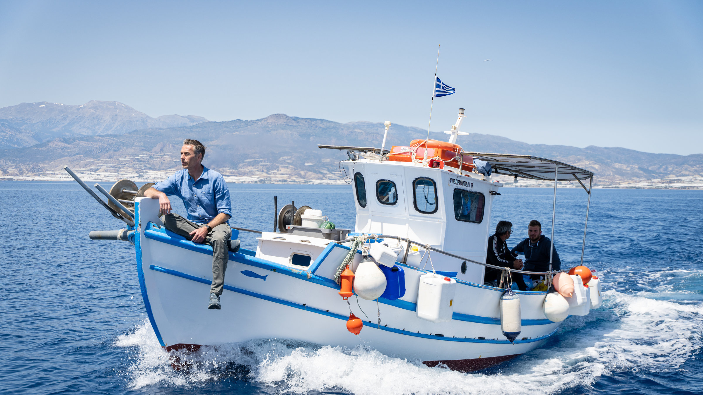 "Terra X: Rätselhafte Phänomene - Powerschnecken und haariges Eis": Moderator Dirk Steffens sitzt auf einem Fischerboot und blickt in Fahrtrichtung auf das Meer vor der Insel Chrysi. Mit an Bord sind zwei weitere Männer.