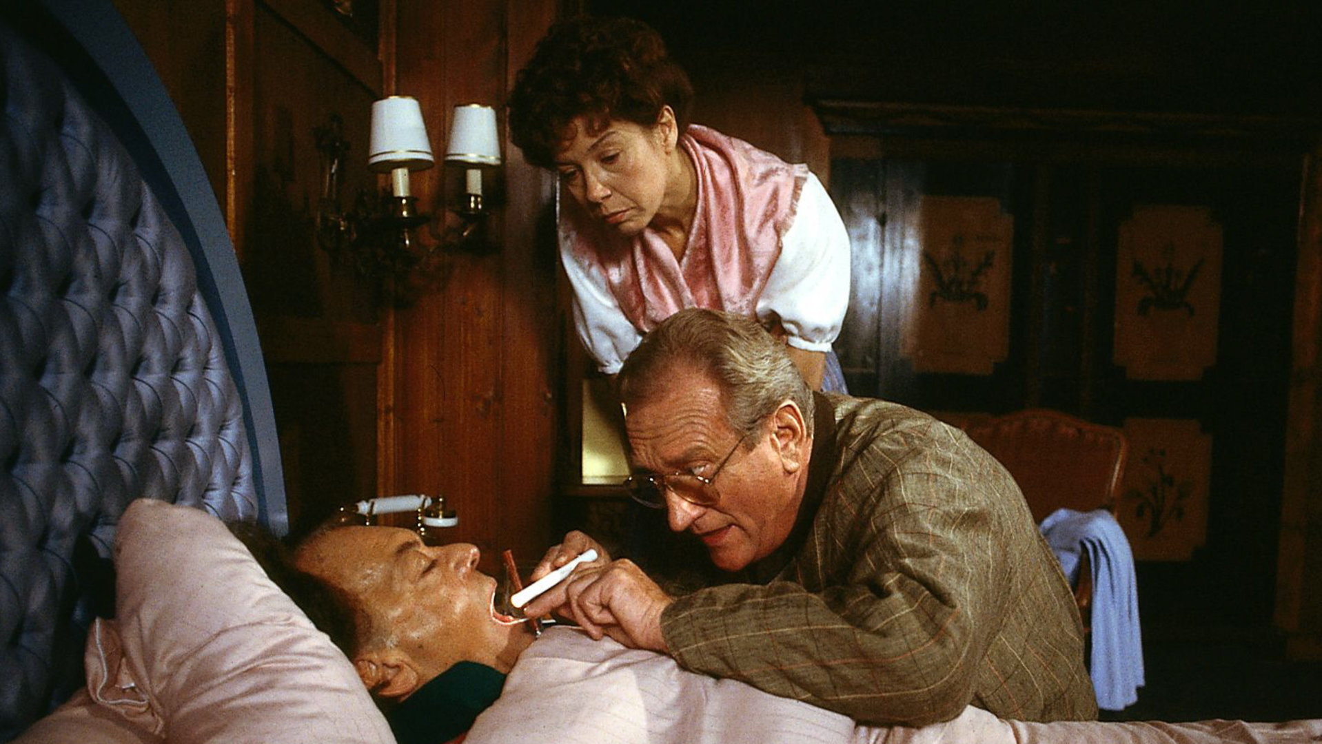 "Die Schwarzwaldklinik - Der kranke Professor": Prof. Brinkmann (Klausjürgen Wussow) liegt im Bett. Dr. Neubert (Dieter Eppler) leuchtet ihm in den Hals, Frau Michaelis (Evelyn Hamann) steht daneben und schaut interessiert zu.