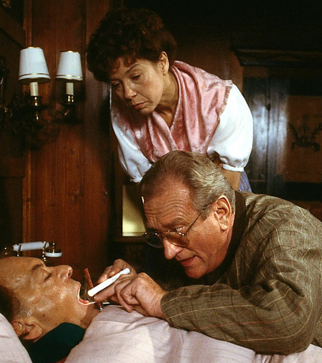 "Die Schwarzwaldklinik - Der kranke Professor": Prof. Brinkmann (Klausjürgen Wussow) liegt im Bett. Dr. Neubert (Dieter Eppler) leuchtet ihm in den Hals, Frau Michaelis (Evelyn Hamann) steht daneben und schaut interessiert zu.