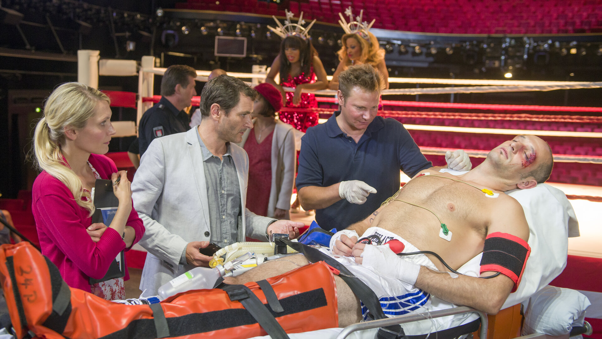 "Notruf Hafenkante - Kampf der Herzen": Der Hauptdarsteller des Musicals "Rocky" (Drew Sarich)  liegt verletzt auf einer Bahre. Dr. Philipp Haase (Fabian Harloff) kümmert sich um ihn. Der künstlerische Leiter Mikkelsen (Alex Brendemühl) und seine Assistentin (Mareile Moeller) stehen dabei und schauen zu. Im Hintergrund ist ein Boxring im Operettenhaus aufgebaut. Zwei Frauen in roten Glitzerkleidern lehnen auf den Ringseilen.