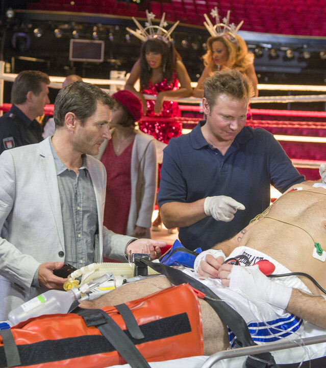 "Notruf Hafenkante - Kampf der Herzen": Der Hauptdarsteller des Musicals "Rocky" (Drew Sarich)  liegt verletzt auf einer Bahre. Dr. Philipp Haase (Fabian Harloff) kümmert sich um ihn. Der künstlerische Leiter Mikkelsen (Alex Brendemühl) und seine Assistentin (Mareile Moeller) stehen dabei und schauen zu. Im Hintergrund ist ein Boxring im Operettenhaus aufgebaut. Zwei Frauen in roten Glitzerkleidern lehnen auf den Ringseilen.