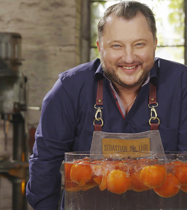 "Die Tricks der Lebensmittelindustrie - Hackfleisch, Wackelpudding & Co.":  Sebastian Lege steht mit einem wassergefülltem Glasgefäß mit Tomaten in einer Werkstatt und lächelt in die Kamera.