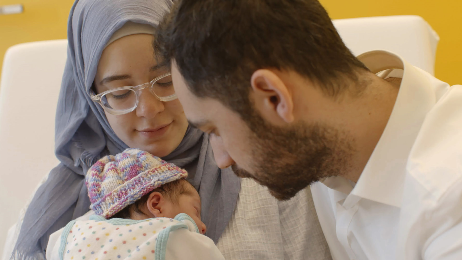 "37°: Guter Hoffnung - Eltern werden in drei Religionen": Ein junges Paar schaut zärtlich auf sein kleines Baby. Die Frau hält das Baby in den Armen, sie trägt Brille und Kopftuch. Das Baby eine bunte Wollmütze.