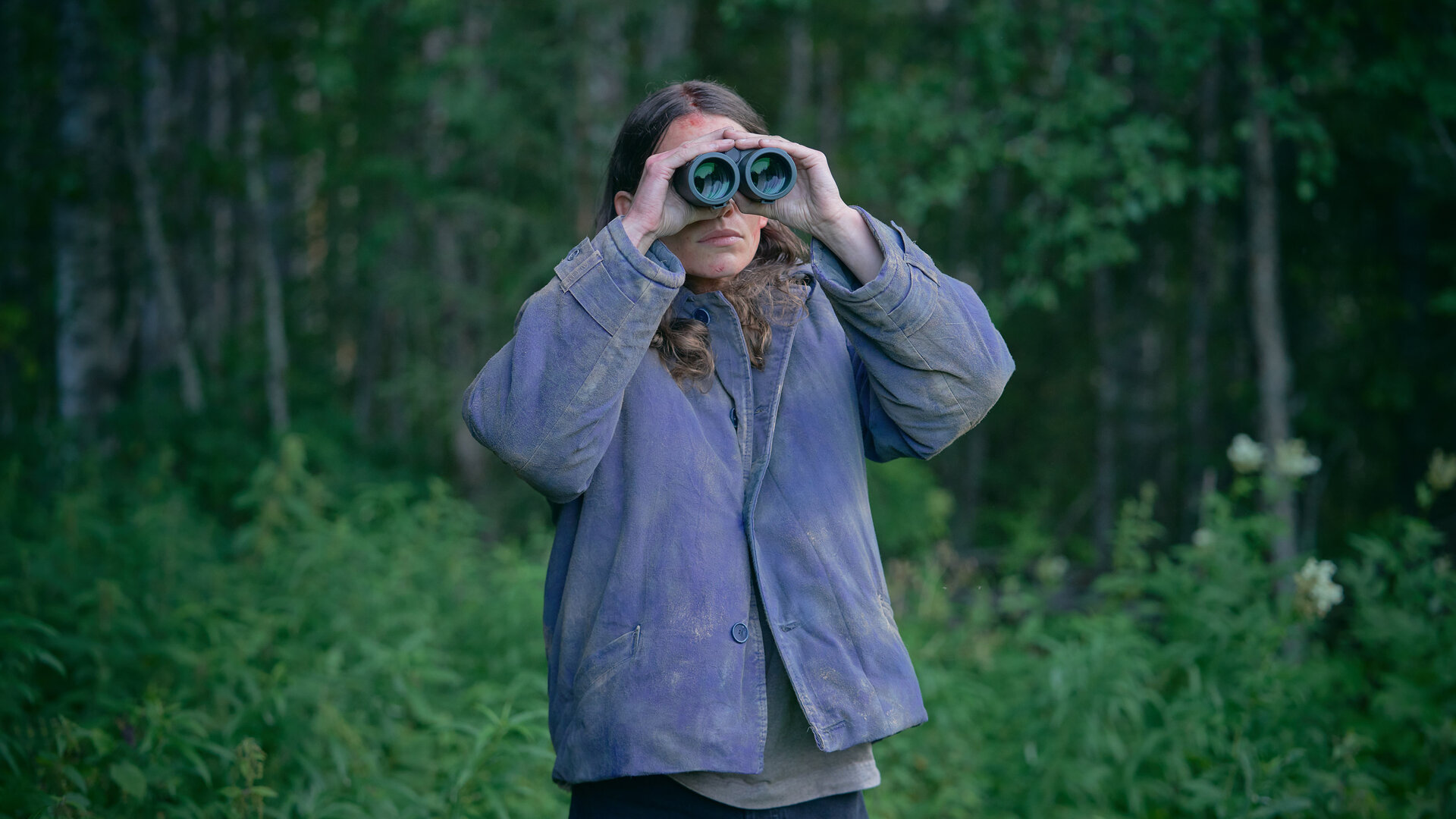 "Wolfssommer – Blutige Spuren - Eine schlechte Entscheidung": Kat (Eliot Sumner) steht vor einem Waldstück. Sie beobachtet etwas durch ein Fernglas.