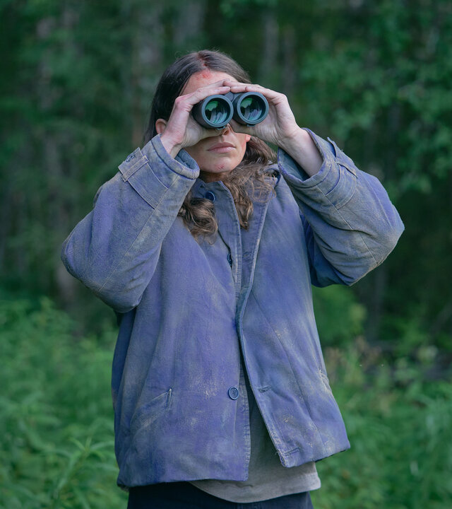 "Wolfssommer – Blutige Spuren - Eine schlechte Entscheidung": Kat (Eliot Sumner) steht vor einem Waldstück. Sie beobachtet etwas durch ein Fernglas.