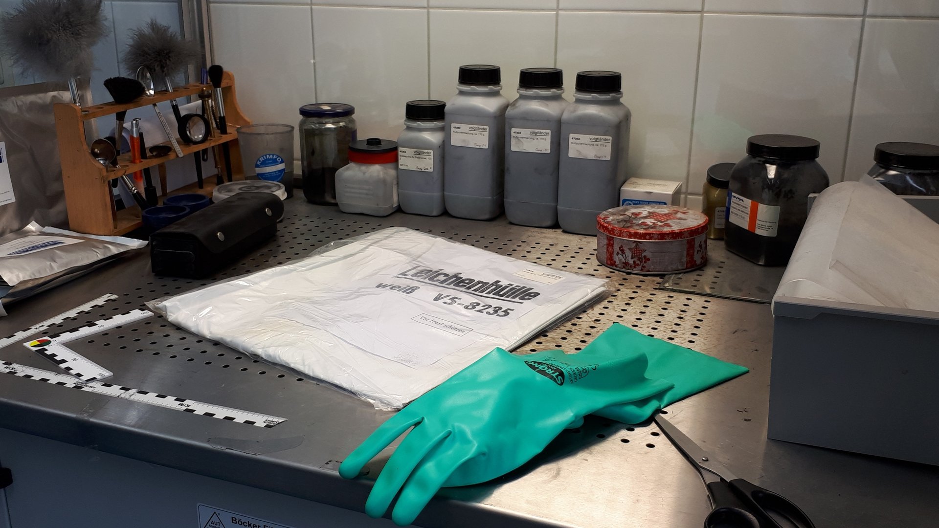 "37°: Die Kriminalisten - Dem Verbrechen auf der Spur": Auf einem Labortisch liegen ein eingepackter, weißer Sack mit der Aufschrift "Leichenhülle", davor ein grüner Gummihandschuh und eine Schere. Im Hintergrund sind Maßband, Pinsel, Lupe und weitere Utensilien zur Spurensicherung zu sehen.