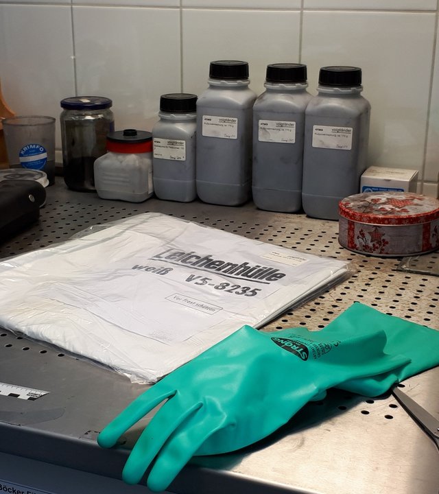 "37°: Die Kriminalisten - Dem Verbrechen auf der Spur": Auf einem Labortisch liegen ein eingepackter, weißer Sack mit der Aufschrift "Leichenhülle", davor ein grüner Gummihandschuh und eine Schere. Im Hintergrund sind Maßband, Pinsel, Lupe und weitere Utensilien zur Spurensicherung zu sehen.