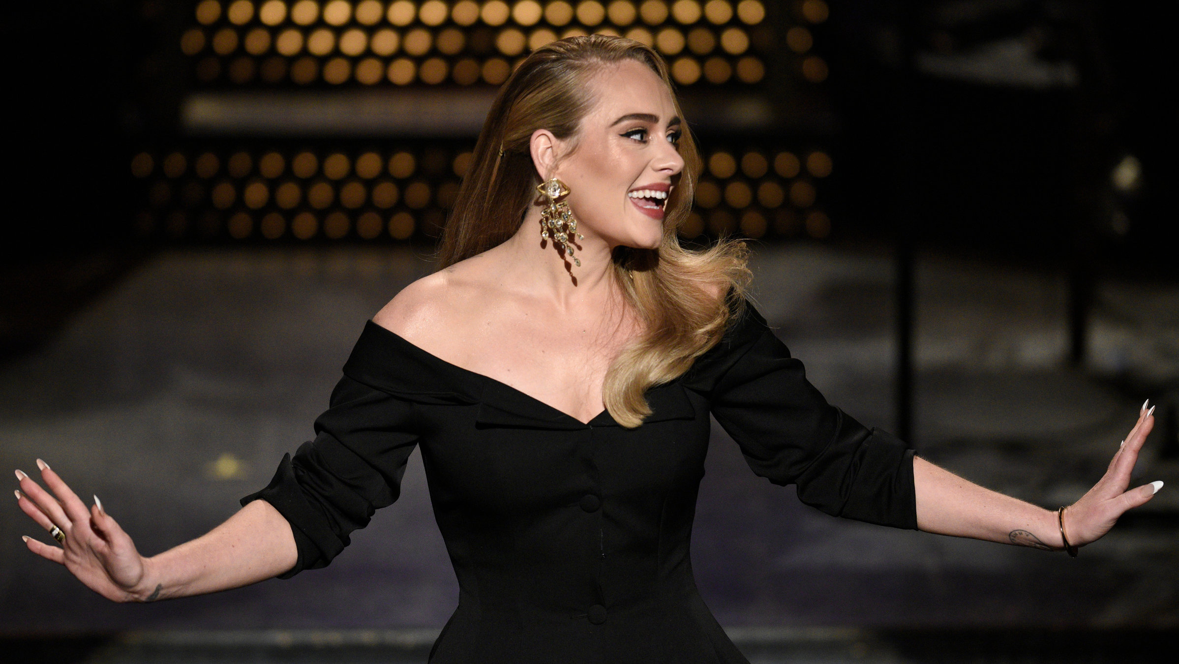 "The True Story of Adele": Adele mit offenem Haar, einem schwarzen Kleid und großen Goldohrringen steht auf einer Bühne, schaut nach rechts und lächelt. Sie streckt die Arme aus.