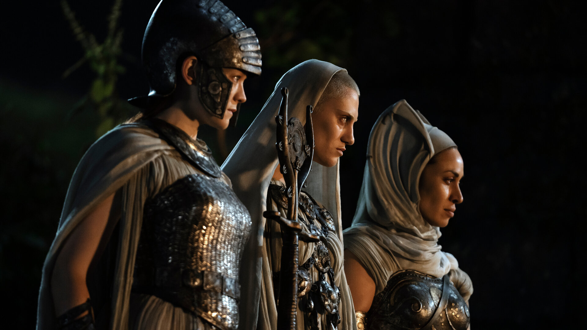 "Der Herr der Ringe: Die Ringe der Macht - Das Auge": Die drei Mystikerinnen (Edith Poor, Bridie Sisson, Kali Kopae) stehen nebeneinander im Dunkeln und beobachten etwas. Eine von ihnen trägt einen Helm, die anderen beiden Kopftücher.