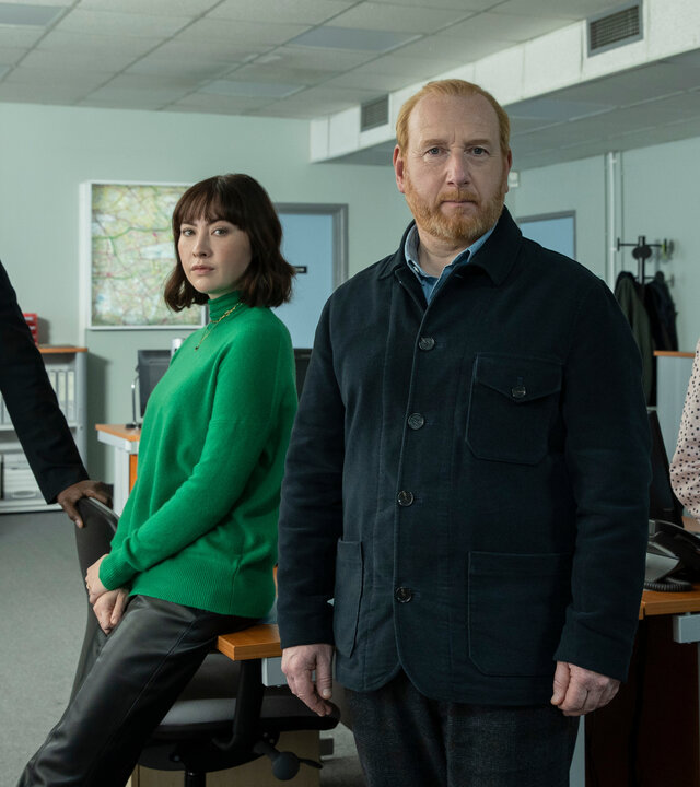 "The Chelsea Detective - Golden Years": DS Layla Walsh (Vanessa Emme) und DC Jess Lombard (Lucy Phelps) lehnen sich im Büro an einen Schreibtisch. Daneben steht DC Connor Pollock (Peter Bankolé), im Vordergrund DI Max Arnold (Adrian Scarborough).