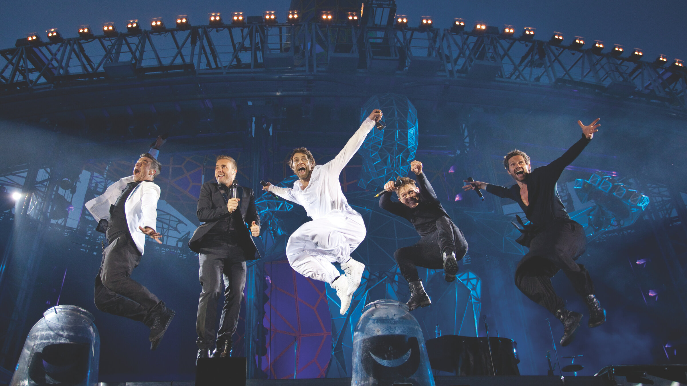 "Take That: Progress Live:" Die Mitglieder der Band Take That während eines Konzerts auf der Bühne. Sie springen alle in die Luft und strecken zum Teil die Arme in die Höhe. Gekleidet sind alle Sänger nur in den Farben Schwarz oder Weiß.