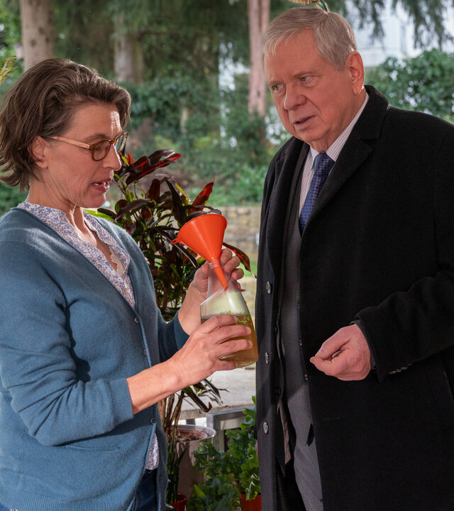 "Der Staatsanwalt - Der Tod hat Vorfahrt": Johanna Will (Jule Gartzke) und Bernd Reuther (Rainer Hunold) stehen in einem hellen Raum vor einem großen Fenster mit Blick ins Grüne. Hinter ihnen stehen viele Topfpflanzen auf der Fensterbank. Johanna Will schaut auf ein Gefäß mit gelber Flüssigkeit und einen orangenen Trichter, die sie in ihren Händen hält. Reuther sieht Will skeptisch an.