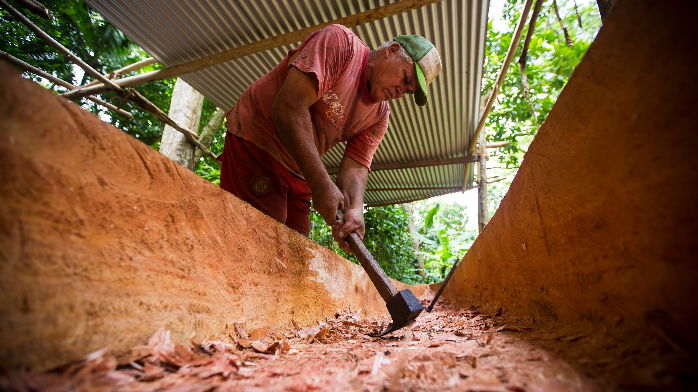 "Schutzprojekt Erde": Aus dem Rumpf einer im Bau befindlichen Piroge aufgenommen:  aus einem einzigen Baumstamm hackt ein Mann in verschmutzter roter Kleidung und Base-Cap Holz aus dem Stamm. Er arbeitet im Freien, geschützt nur durch ein Wellblechdach auf einer Konstruktion aus Holzstangen.