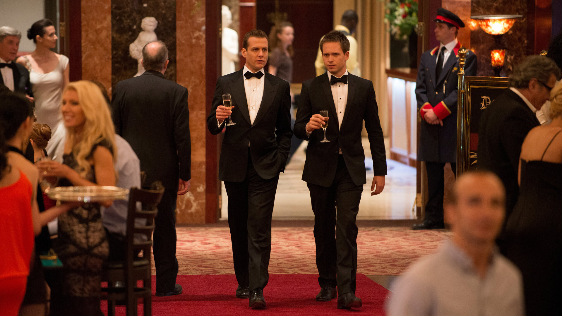 "Suits - Volles Risiko": Im Casino: Harvey (Gabriel Macht) und Mike (Patrick J. Adams) schreiten im Smoking über den roten Teppich, mit jeweils einem Sektglas in der Hand. Um sie herum herrscht geschäftiges Treiben in glamouröser Kulisse.