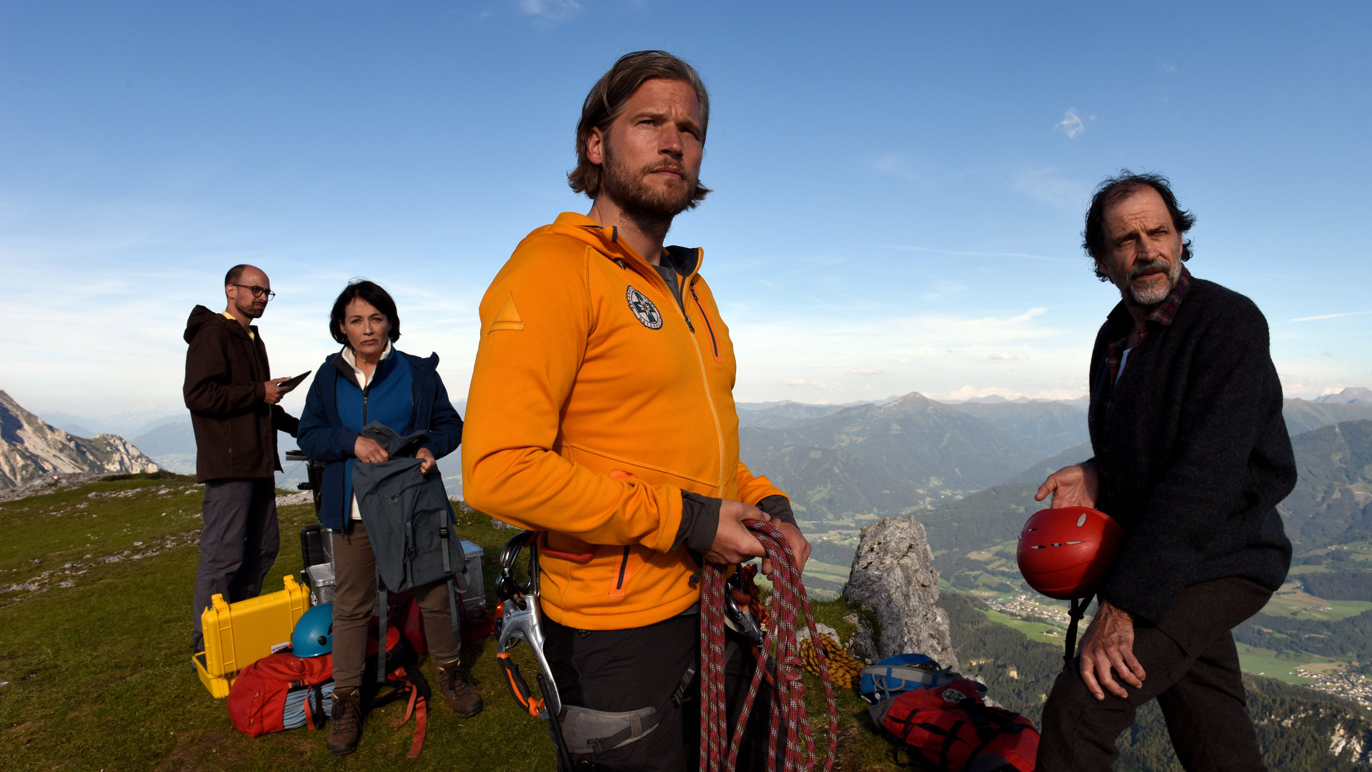 "Die Bergretter - Altes Eisen": Auf einer Bergspitze mit Blick ins Tal stehen der Bergretter Markus (Sebastian Ströbel) und Walther Senn (Helmfried von Lüttichau) und blicken in die Ferne. Im Hintergrund stehen Tabea Gollini (Anke Sevenich) und Yasin Bardak (Roman Blumenschein) zwischen diversen Kisten und Taschen.