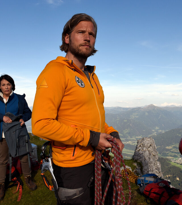 "Die Bergretter - Altes Eisen": Auf einer Bergspitze mit Blick ins Tal stehen der Bergretter Markus (Sebastian Ströbel) und Walther Senn (Helmfried von Lüttichau) und blicken in die Ferne. Im Hintergrund stehen Tabea Gollini (Anke Sevenich) und Yasin Bardak (Roman Blumenschein) zwischen diversen Kisten und Taschen.
