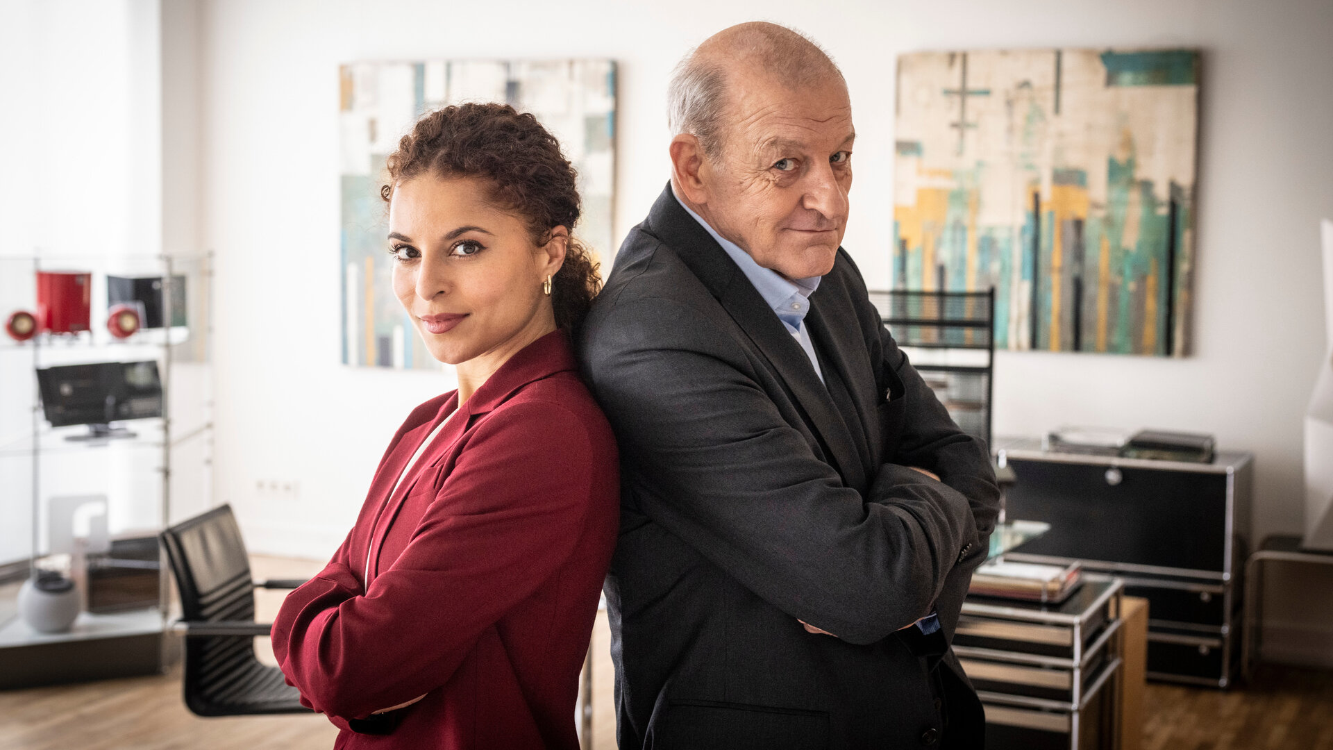 "Wilsberg – Trügerische Sicherheit": Dr. Tessa Tilker (Patricia Meeden) steht Rücken an Rücken zu Georg Wilsberg (Leonard Lansink) in einem Büro. Beide verschränken die Arme und schauen mit einem entschlossenen Blick in die Kamera.