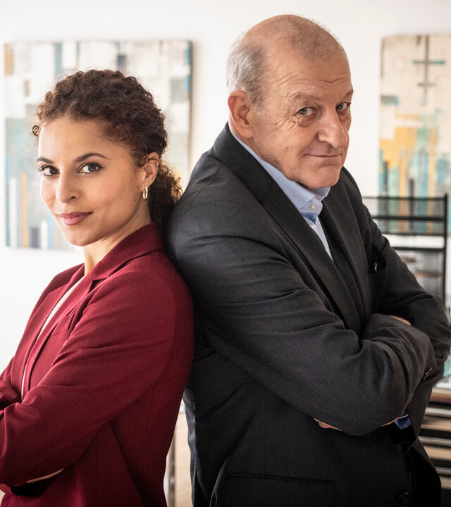 "Wilsberg – Trügerische Sicherheit": Dr. Tessa Tilker (Patricia Meeden) steht Rücken an Rücken zu Georg Wilsberg (Leonard Lansink) in einem Büro. Beide verschränken die Arme und schauen mit einem entschlossenen Blick in die Kamera.