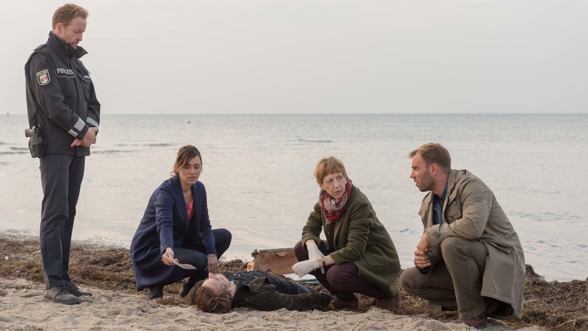 "SOKO Wismar – Luftbild": Kai Timmermann (Mathias Junge) steht am Strand und sieht zur Leiche von Marcel Jeske (Benjamin Trinks), die vor ihm im Sand liegt. Karoline Joost (Nike Fuhrmann),  Rechtsmedizinerin Helene Sturbeck (Katharina Blaschke) und Lars Pöhlmann (Dominic Boeer) haben sich um die Leiche versammelt und unterhalten sich.