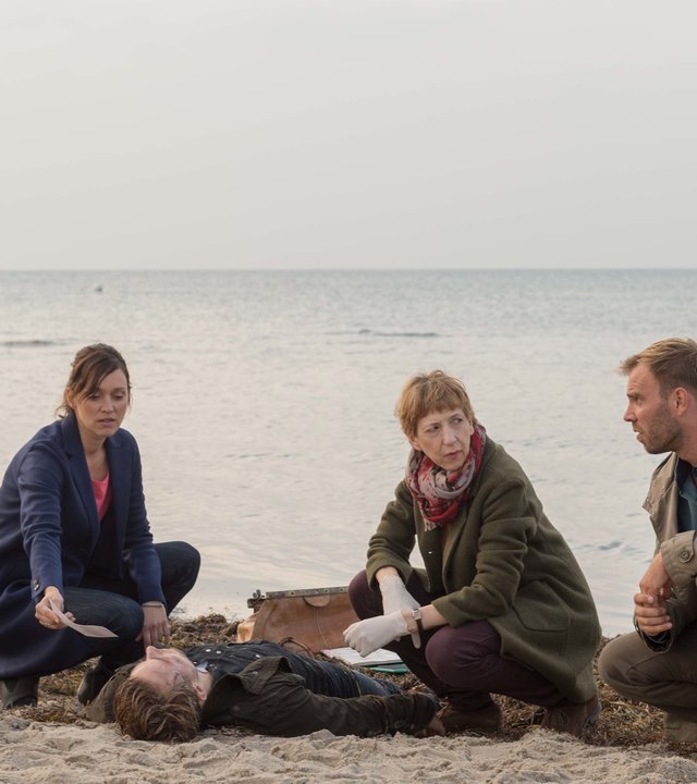 "SOKO Wismar – Luftbild": Kai Timmermann (Mathias Junge) steht am Strand und sieht zur Leiche von Marcel Jeske (Benjamin Trinks), die vor ihm im Sand liegt. Karoline Joost (Nike Fuhrmann),  Rechtsmedizinerin Helene Sturbeck (Katharina Blaschke) und Lars Pöhlmann (Dominic Boeer) haben sich um die Leiche versammelt und unterhalten sich.