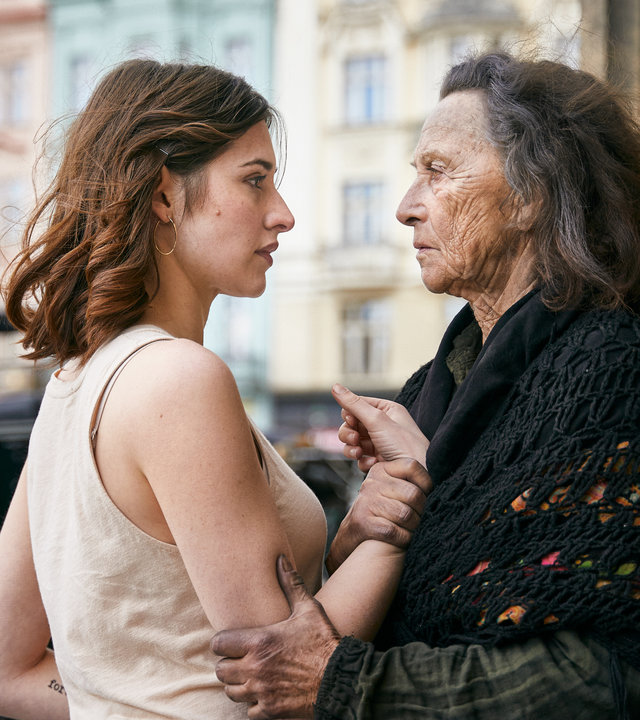 "Der Schatten - Die Prophezeiung": Norah Richter (Deleila Piasko) wird von der Bettlerin Dorothea Lechner (Elisabeth Rath) fest am Arm gepackt. Die beiden stehen sich gegenüber und schauen sich eindringlich in die Augen. Im Hintergrund sieht man eine Kutsche vor einer Hausfassade.