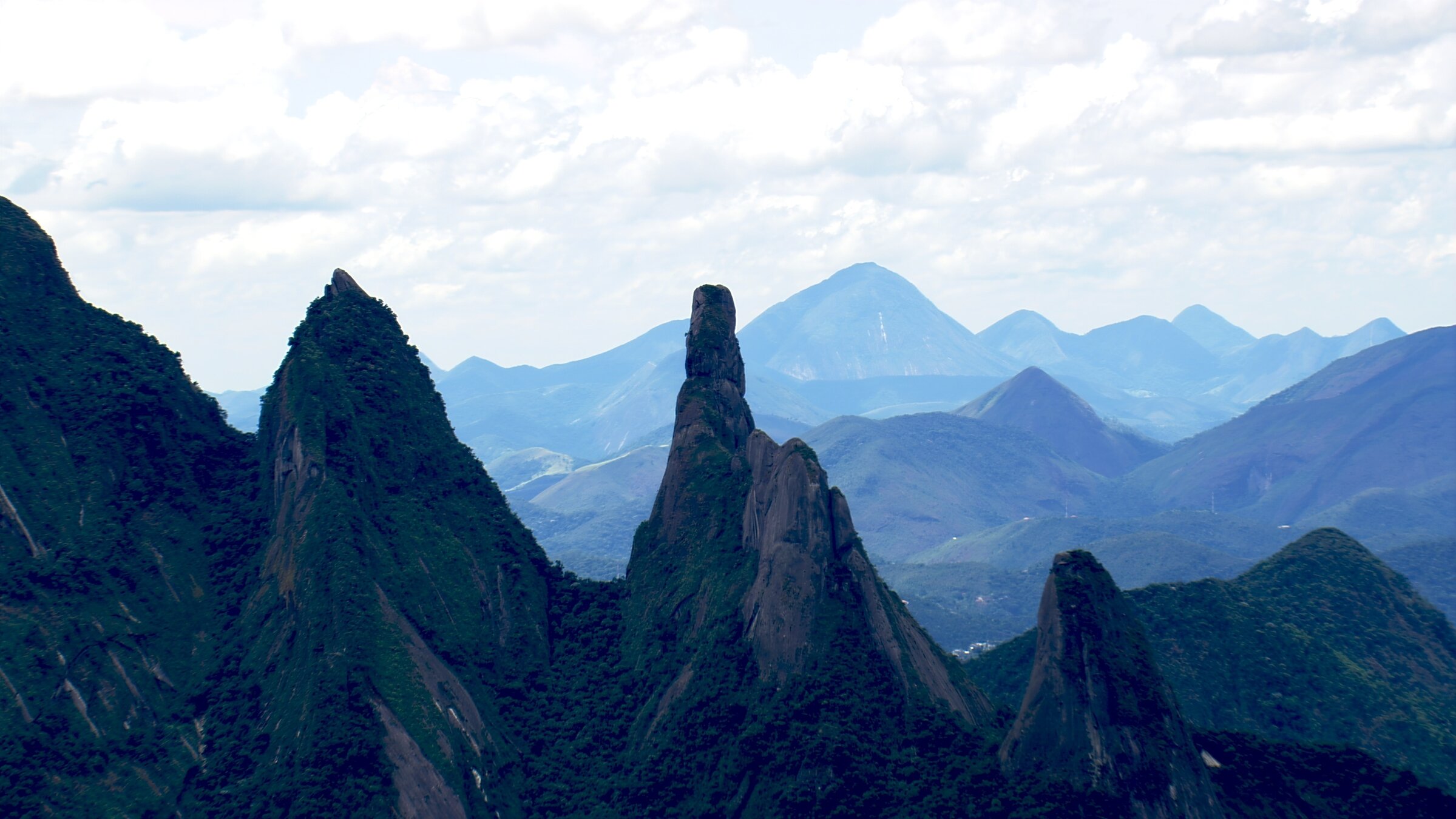 "Im Flug über Rio de Janeiro": Bizarre Felsformationen vor einer bewaldeten Berglandschaft, im Hintergrund sind Wolken.