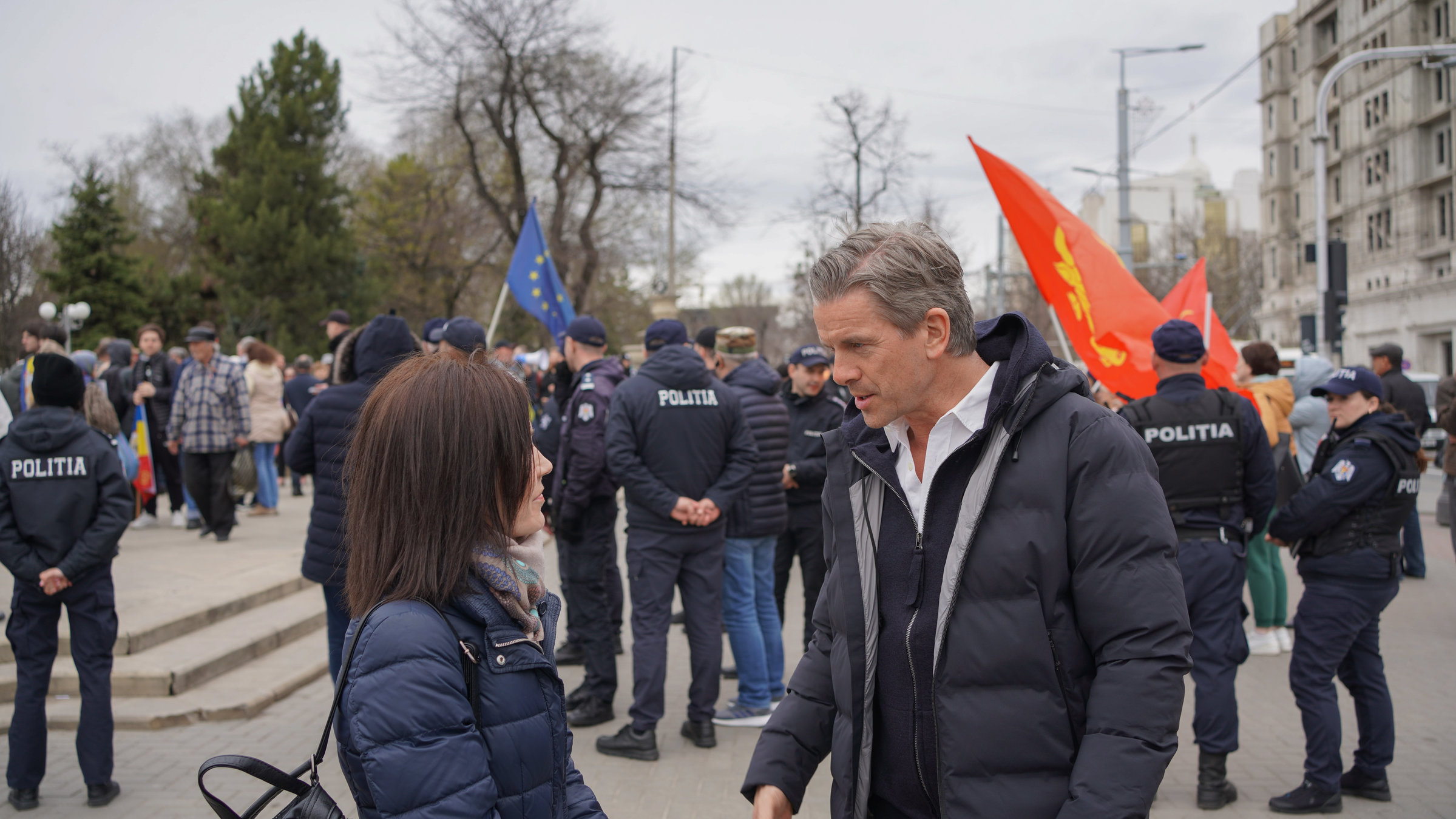 "Markus Lanz – Moldawien ungeschminkt": Markus Lanz und eine Frau stehen auf einer Straße und unterhalten sich. Im Hintergrund ist eine Menschenmenge zu sehen, an deren Rand Polizisten stehen.