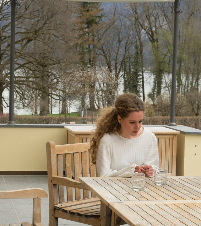 "Der Alte - Schmutzige Wäsche": Caro Schaller (Klara Deutschmann) sitzt an einem Tisch, auf dem Gläser und eine Karaffe mit Wasser stehen. Richard Voss (Jan-Gregor Kremp) steht rechts daneben und schaut sie an. Im Hintergrund ist Annabell Lorenz (Stephanie Stumph), sie steht mit dem Rücken zu ihnen hat aber den Kopf in Richtung Tisch gedreht. Sie stehen auf einer Terrasse, dahinter sieht man einen See und Berge.