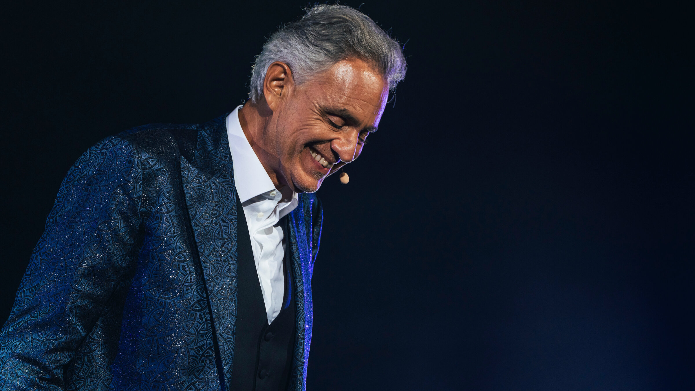 "Andrea Bocelli: Romanza": Das Bild zeigt eine einzelne Person auf einer Bühne vor einem dunklen, nahezu schwarzen Hintergrund. Die Szene ist durch gezielte Bühnenbeleuchtung in kühlen Blau- und Violetttönen ausgeleuchtet, die insbesondere die Konturen und die Kleidung hervorheben.