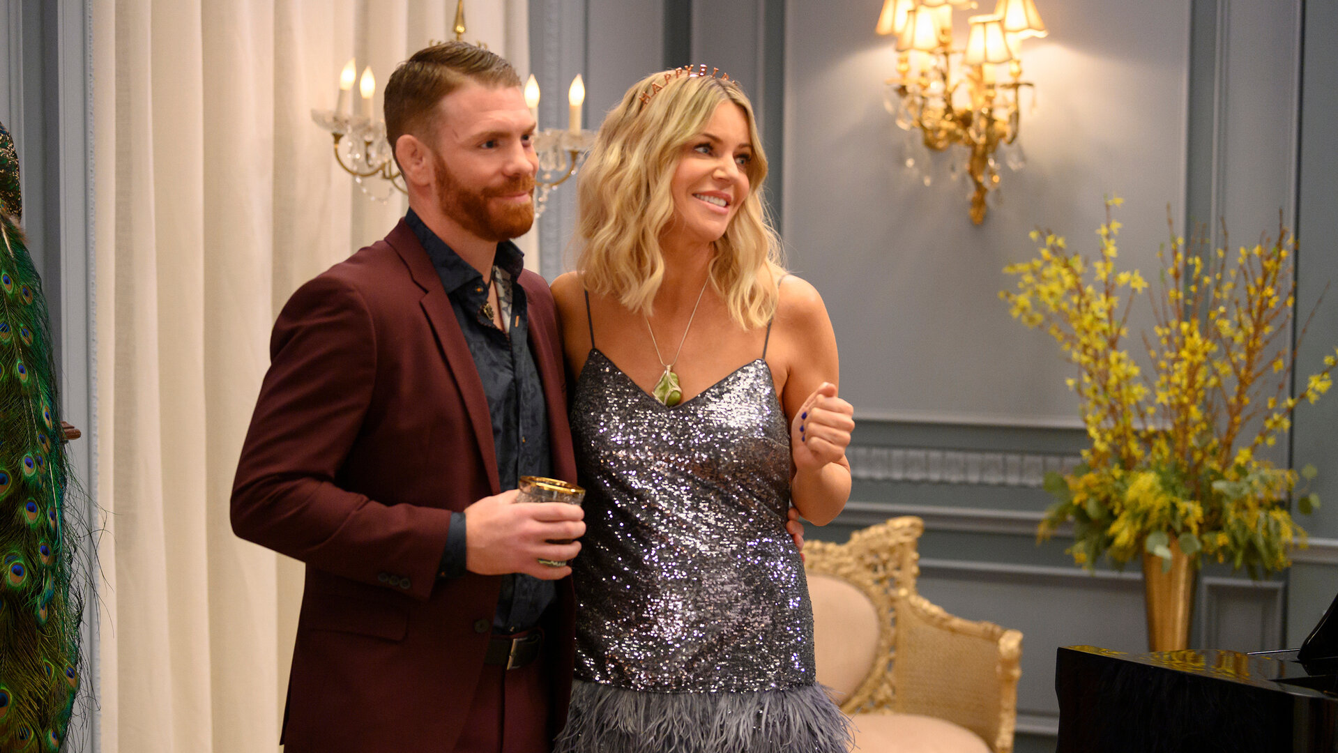 "Hacks - Da ist mehr": DJ (Kaitlin Olson) und Aidan (Paul Felder) stehen eng nebeneinander in einem eleganten Raum mit blauen Wänden, Blumendekor und Kronleuchtern. DJ blickt lächelnd zur Seite, während sie ihren Arm um Aidans Rücken gelegt hat, der entspannt geradeaus schaut.