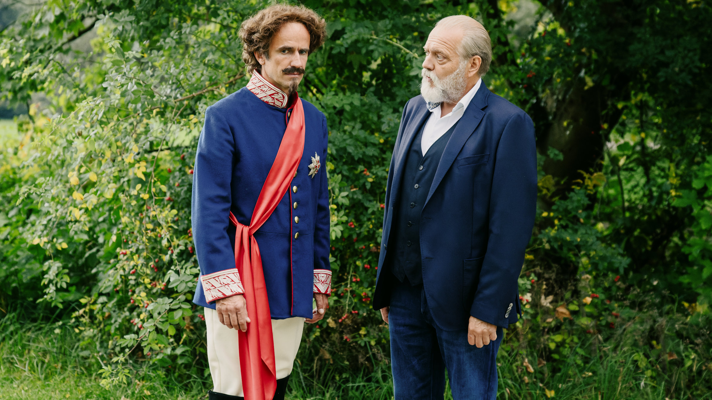 "Der Alte - Der Mondkönig": Robert Obermüller (Felix Hellmann), in der Rolle von Ludwig II. eingekleidet, steht mit Richard Voss (Jan-Gregor Kremp) auf einer Wiese im Park. Die beiden sprechen miteinander. Robert Obermüller dreht sich ertappt weg von Voss.